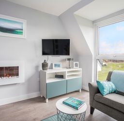 A living room with a wall-mounted TV a fireplace a gray sofa with blue cushions a sideboard and a round coffee table at Isallt in Trearddur Bay