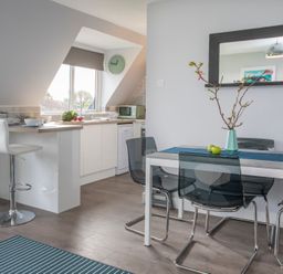 A kitchen and dining area with white cabinets, bar stools, a dining table, chairs, a mirror, and a vase with branches at Isallt Trearddur Bay