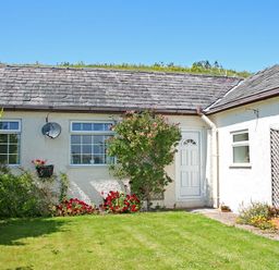 The exterior of a cottage with a grass lawn flowers around the walls and a white door at Mermaid Cottage in Brynsiencyn