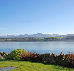 A view of a calm body of water with greenery and rocks in the foreground and hills and houses in the background at Mermaid Cottage in Brynsiencyn