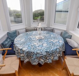 A dining area with a round table covered with a patterned tablecloth surrounded by built-in cushioned seating and wooden chairs at Min Y Mor Trearddur Bay