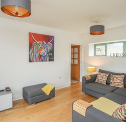 A living room with a gray sectional sofa and colorful cushions a gray ottoman wooden flooring and a colorful cow painting on the wall at Mynydd Parys in Brynsiencyn