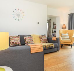 A living room with a gray sofa with colorful pillows a yellow armchair a yellow lamp and a clock on the wall at Mynydd Parys in Brynsiencyn