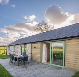 An outdoor patio area with a table and six chairs beside a house with stone and wood siding at Mynydd Parys in Brynsiencyn