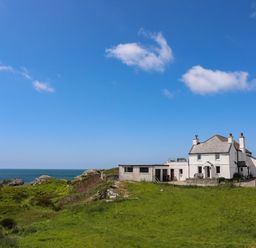 A white house near the coast with green grass and rocks under a blue sky at Pant yr Hyman in Rhoscolyn