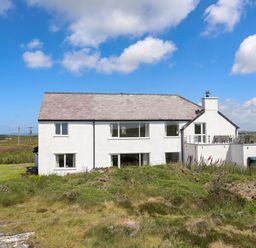 A white two-story house with a slate roof and glass balcony surrounded by grass and shrubs at Pant yr Hyman in Rhoscolyn