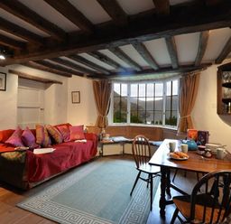 A living room with a red-covered sofa with cushions a wooden dining table with chairs and a bay window with curtains at Pantlleni Nant Gwynant