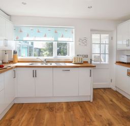 A kitchen with white cabinets wooden countertops a window with a sailboat blind a toaster and cutting board with vegetables at Pencraig in Trearddur Bay