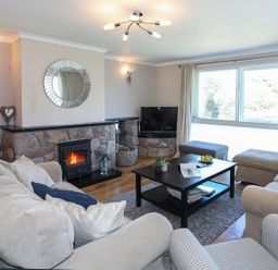 A living room with a stone fireplace a round mirror a television beige sofas a black coffee table and large window at Pencraig in Trearddur Bay