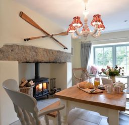 A dining area with a wooden table set with mugs bread and flowers near a wood stove with oars mounted above at Penrhyn Farm in Moelfre
