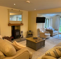 A living room with beige sofas, a coffee table, a wood-burning stove, an armchair, a wall-mounted television, a wicker sofa, and glass doors leading to a garden at Penrhyn Farm in Moelfre