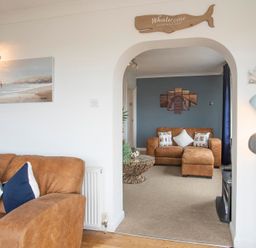 A living room with brown sofas and decorative pillows with a wooden whale sign above an archway at Pen Y Daith in Aberffraw