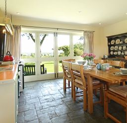 A kitchen dining area with wooden table and chairs set with dishes and flowers near large windows at Perth Hwfa in Malltraeth