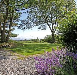 A garden with grass and purple flowers bordered by gravel and trees at Perth Hwfa in Malltraeth