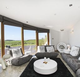 A living room with gray sofas and cushions a round white coffee table black rug and large windows showing green fields at Rhyd Angharad Barns in Rhydwyn