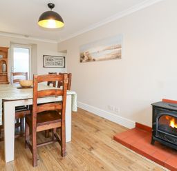 A dining room with a table and wooden chairs a wood burning stove and a wooden cabinet at Seiriol View in Moelfre
