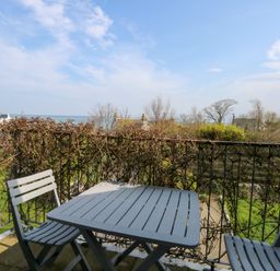 A balcony with two chairs and a table overlooking a garden and houses with the sea in the background at Seiriol View in Moelfre