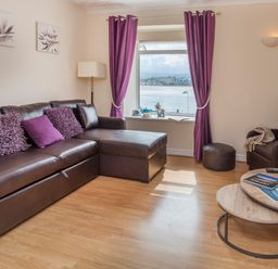A living room with a brown leather sectional sofa with purple cushions a round wooden coffee table with magazines a brown armchair a window with purple curtains and a view of water at The Mermaid in Brynsiencyn