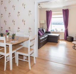 A dining area with a wooden table and white chairs next to a living room with a brown leather sectional sofa and purple curtains at The Mermaid in Brynsiencyn