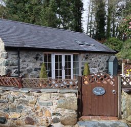 A stone cottage with a wooden gate and fence surrounded by trees and flowers at The Honey Pot in Garndolbenmaen