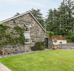 A stone house with climbing plants and a lawn in front surrounded by trees and a paved pathway at The Little Coach House in Garndolbenmaen