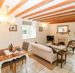 A living room with a sofa a dining table and chairs a fireplace and exposed wooden ceiling beams at The Little Coach House in Garndolbenmaen