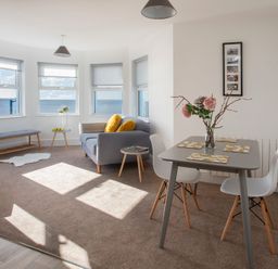 A living room with a gray sofa and yellow cushions a small dining table with two white chairs flowers in a vase a bench and windows showing the sea at The Towers - Ardudwy in Criccieth
