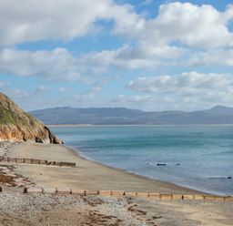 A beach with a rocky cliff on the left and wooden groynes along the shore with mountains in the background at The Towers - Ardudwy in Criccieth