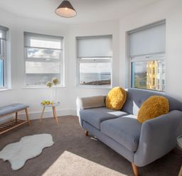 A sitting room with a grey sofa and yellow cushions by windows showing an ocean view at The Towers - Ardudwy in Criccieth