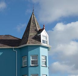 A blue building with a pointed turret and white windows against a cloudy sky at The Towers - Castell in Criccieth
