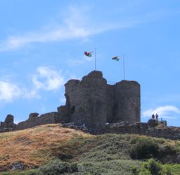An old stone castle on a hill with two flags flying and people standing nearby at The Towers - Llyn in Criccieth