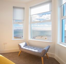 A corner room with three windows overlooking the sea and a gray bench with wooden legs at The Towers - Llyn in Criccieth