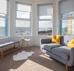 A living room with a gray sofa and yellow cushions near windows overlooking the sea at The Towers - Llyn in Criccieth