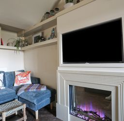 A corner of a living room with a blue sectional sofa with two decorative pillows a small woven stool a built in electric fireplace and a mounted flat screen TV with shelves with plants and framed pictures at Towyn Lodge in Trearddur Bay
