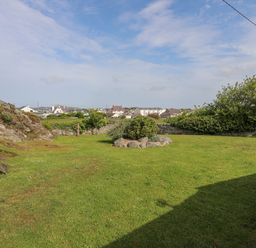 A grassy backyard with shrubs rocks and a stone wall with houses in the background in Trearddur Bay