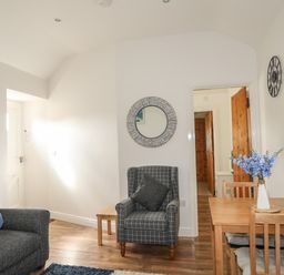 A living room with a gray armchair a wooden table with chairs and flowers and a sofa at Tyn Towyn - Bwthyn Carreg in Trearddur Bay