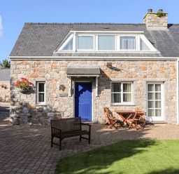 Stone house with blue door wooden bench and table with chairs on paved patio at Tyn Towyn - Ty Glas in Trearddur Bay