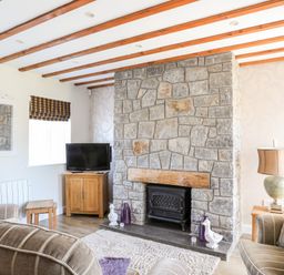 A living room with a stone fireplace wooden beams on ceiling and a television on a wooden cabinet at Tyn Towyn - Ty Glas in Trearddur Bay