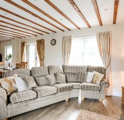 A living room with a striped sectional sofa and cushions near windows with curtains and wooden beams on the ceiling at Tyn Towyn - Ty Oren in Trearddur Bay