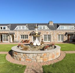 A stone building with multiple windows and a courtyard featuring a circular stone fountain with swan statues in front of it at Tyn Towyn - Ty Piws in Trearddur Bay