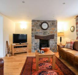 A living room with a leather sofa a stone fireplace a wooden coffee table and a television at Tyn Towyn - The Stables in Trearddur Bay