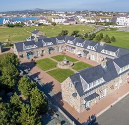 A courtyard with a fountain surrounded by stone buildings with dormer windows and a view of houses and fields at Tyn Towyn - The Stables in Trearddur Bay