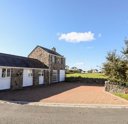A stone building with white doors and windows next to a paved driveway and trees at Tyn Towyn - The Stables in Trearddur Bay