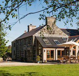 A stone house with outdoor wooden table and chairs under an umbrella in a garden at Tyddyn Morthwyl in Criccieth