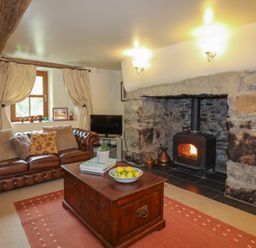 A living room with a leather sofa wooden coffee table with books and fruit bowl and a stone fireplace with a wood burning stove at Tyddyn Morthwyl in Criccieth