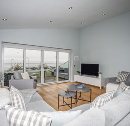 A living room with grey couches and checkered pillows and a TV on a stand near large glass sliding doors at Ty Glyn in Criccieth