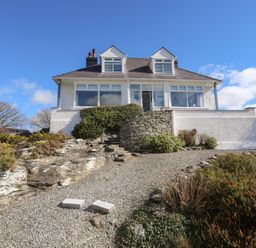 A white house with dormer windows and stone steps surrounded by bushes and gravel path at Ty Llwyd in Trearddur Bay