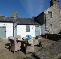 An outdoor patio area with a round glass table and six wicker chairs in front of a stone and whitewashed house at Ty Mawr Porthmadog in Porthmadog