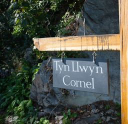 A wooden post with a hanging sign reading Tyn Llwyn Cornel surrounded by rocks and plants at Tyn Llwyn Cornel in Beddgelert
