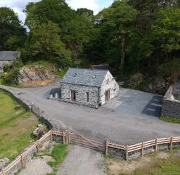A stone cottage with a slate roof surrounded by trees and gravel driveway at Tyn Llwyn Cornel in Beddgelert